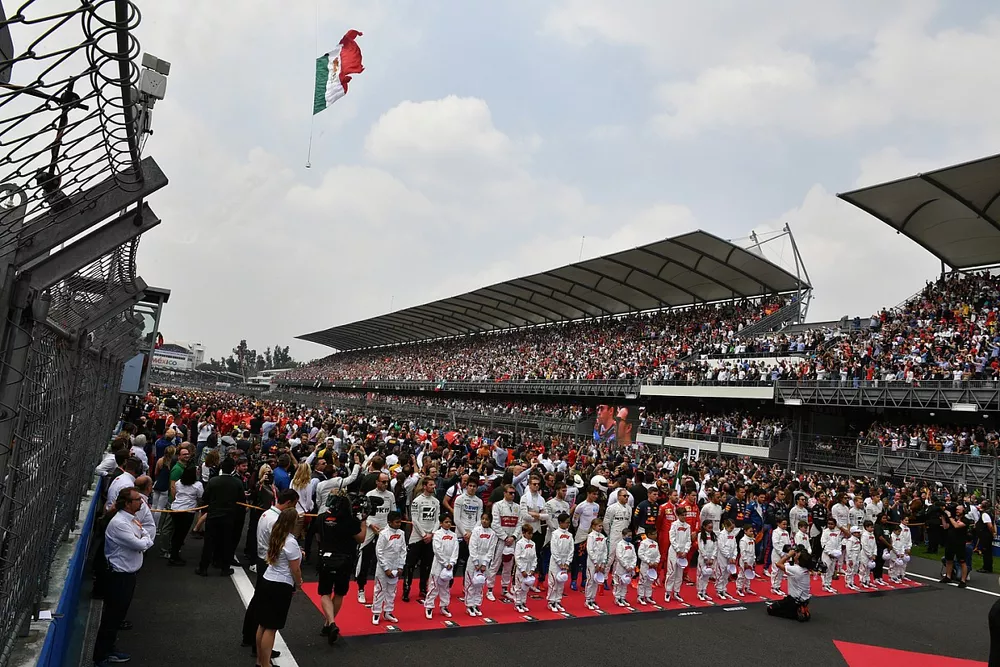 Los pilotos se paran con las mascotas de Grid Kid durante el himno nacional mientras un helic&oacute;ptero ondea la bandera antes de la salida